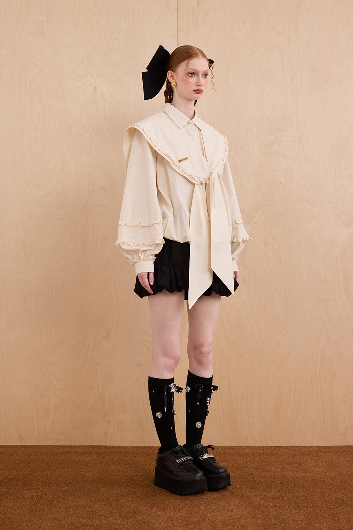 Model wearing a cream blouse with ruffled details, black skirt, and black shoes against a beige background