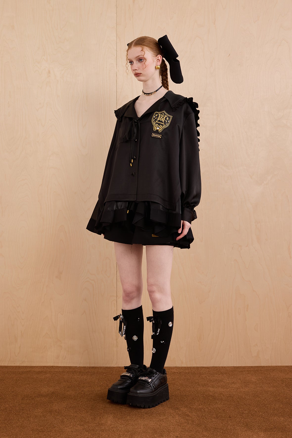Model wearing a black outfit with a high collar and skirt, standing against a beige wall.
